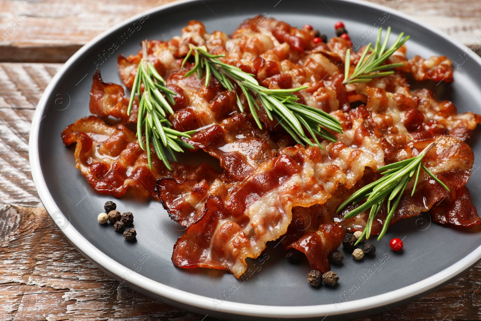 Photo of Slices of tasty fried bacon, peppercorns and rosemary on wooden table, closeup