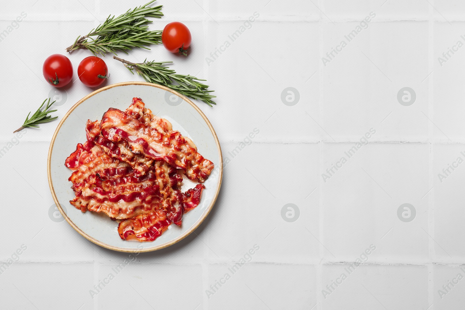 Photo of Slices of tasty fried bacon, rosemary and tomatoes on white tiled table, top view. Space for text