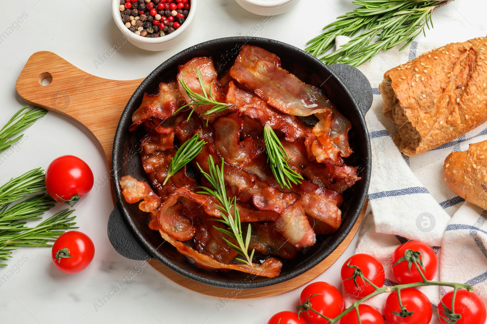 Photo of Delicious bacon slices in frying pan, spices and tomatoes on white table, flat lay