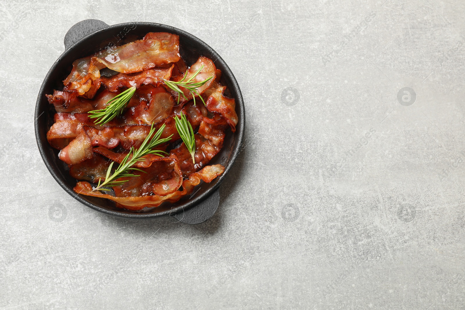 Photo of Delicious bacon slices and fresh rosemary in frying pan on light grey table, top view. Space for text