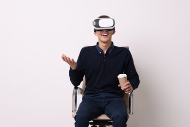 Photo of Happy young man with virtual reality headset and cup of drink sitting on chair near white wall