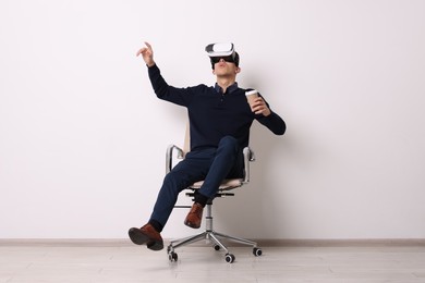 Photo of Emotional young man with virtual reality headset and cup of drink sitting on chair near white wall