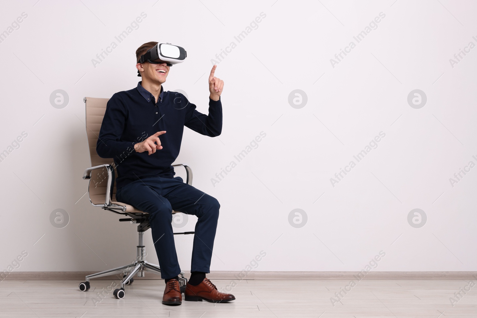 Photo of Happy young man with virtual reality headset sitting on chair near white wall, space for text