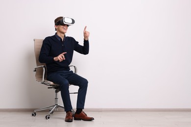 Happy young man with virtual reality headset sitting on chair near white wall, space for text