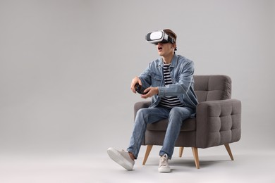 Emotional young man with virtual reality headset and controller sitting on armchair against light grey background, space for text