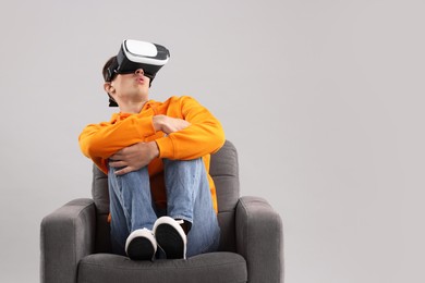 Emotional young man with virtual reality headset sitting on armchair against light grey background, space for text