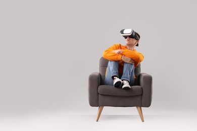 Emotional young man with virtual reality headset sitting on armchair against light grey background, space for text