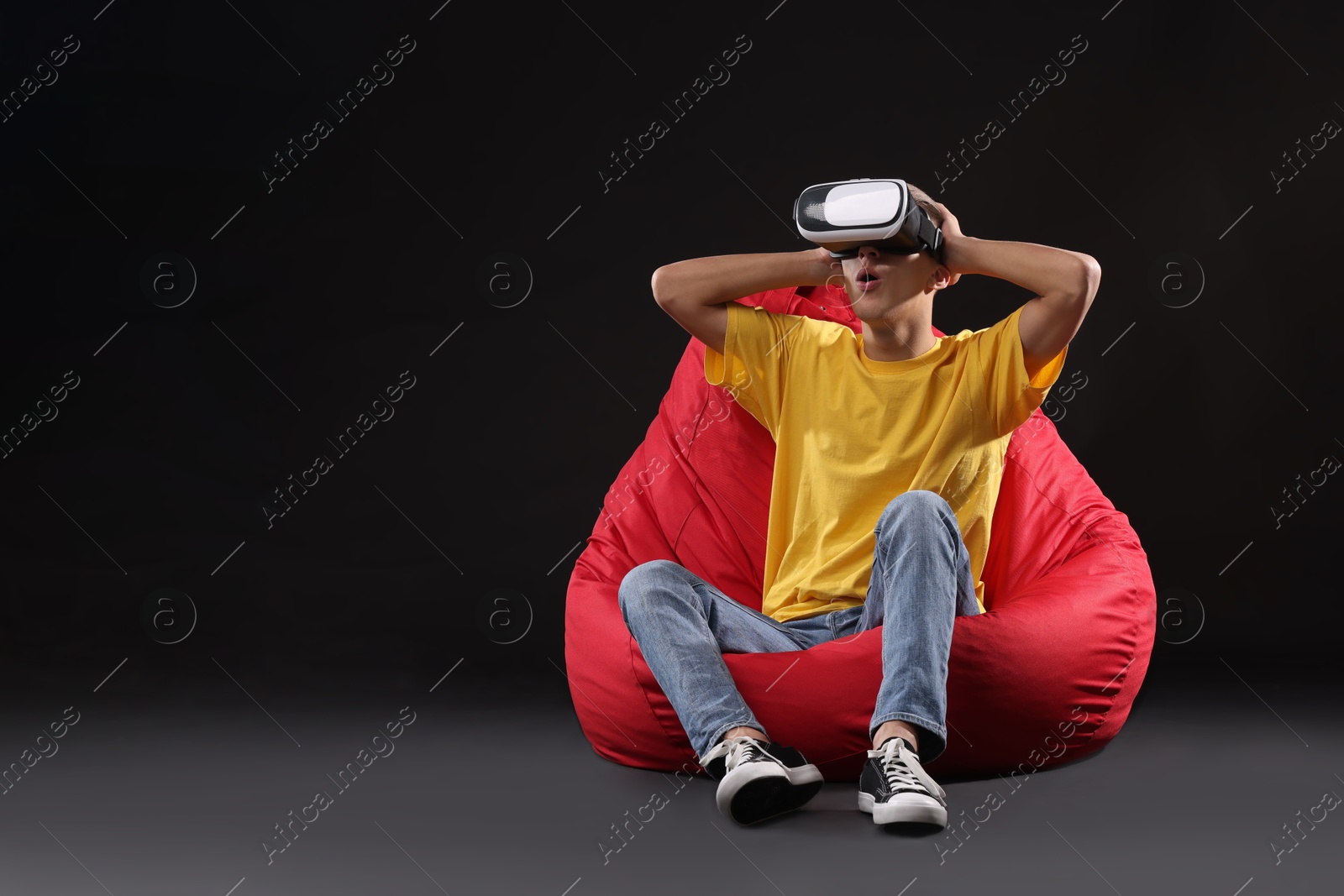 Photo of Emotional young man with virtual reality headset sitting on bean bag chair against black background, space for text