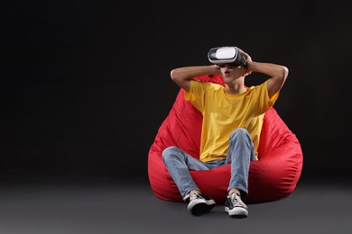 Emotional young man with virtual reality headset sitting on bean bag chair against black background, space for text