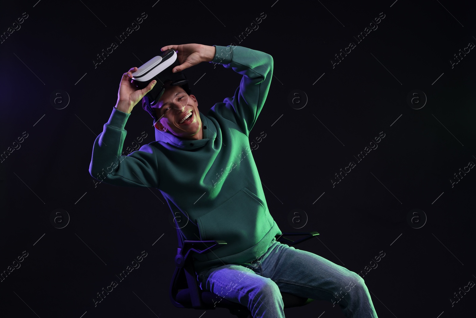 Photo of Happy young man with virtual reality headset sitting on chair in neon lights against black background