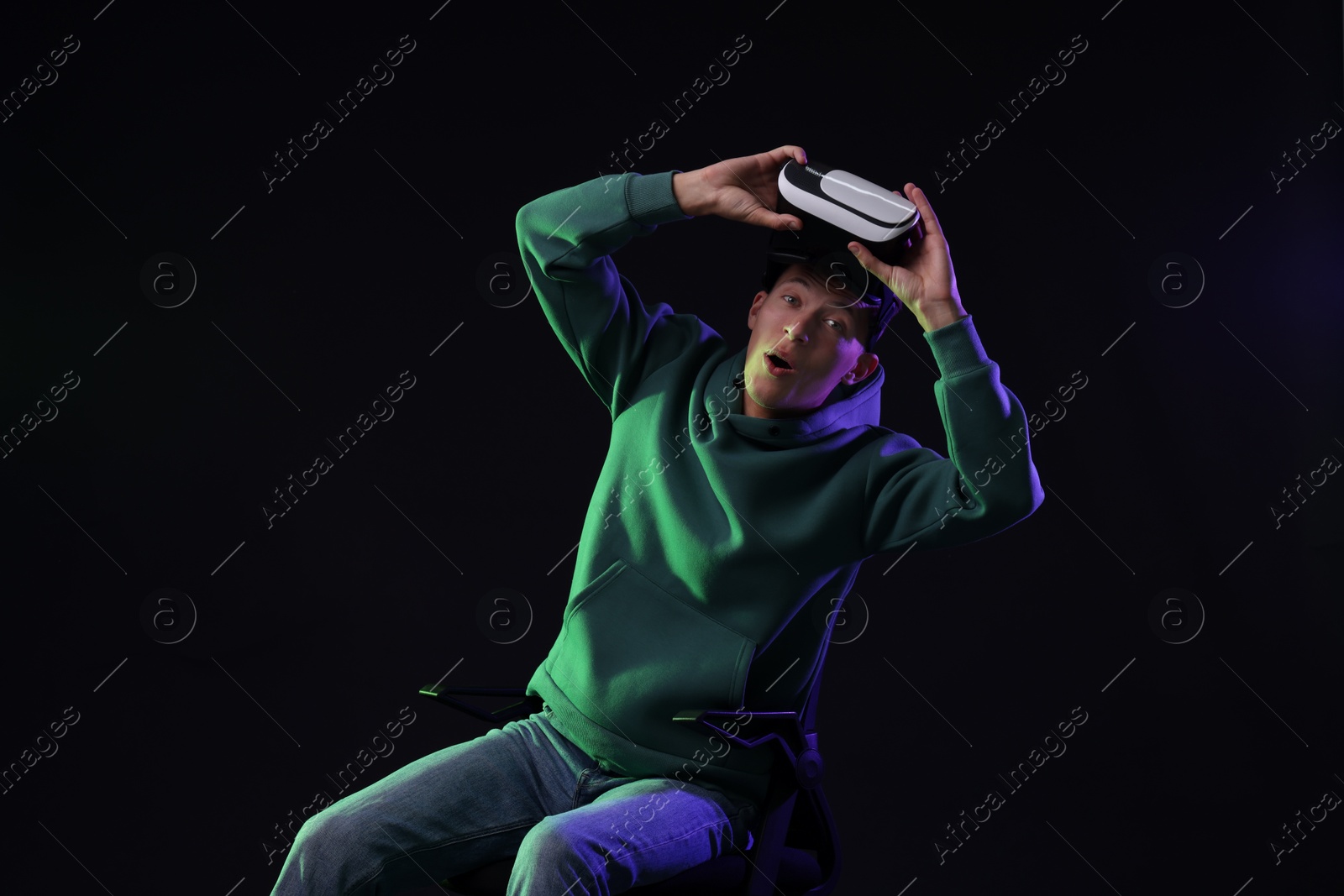 Photo of Emotional young man with virtual reality headset sitting on chair in neon lights against black background