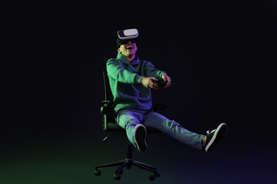 Photo of Happy young man with virtual reality headset and controller sitting on chair in neon lights against black background