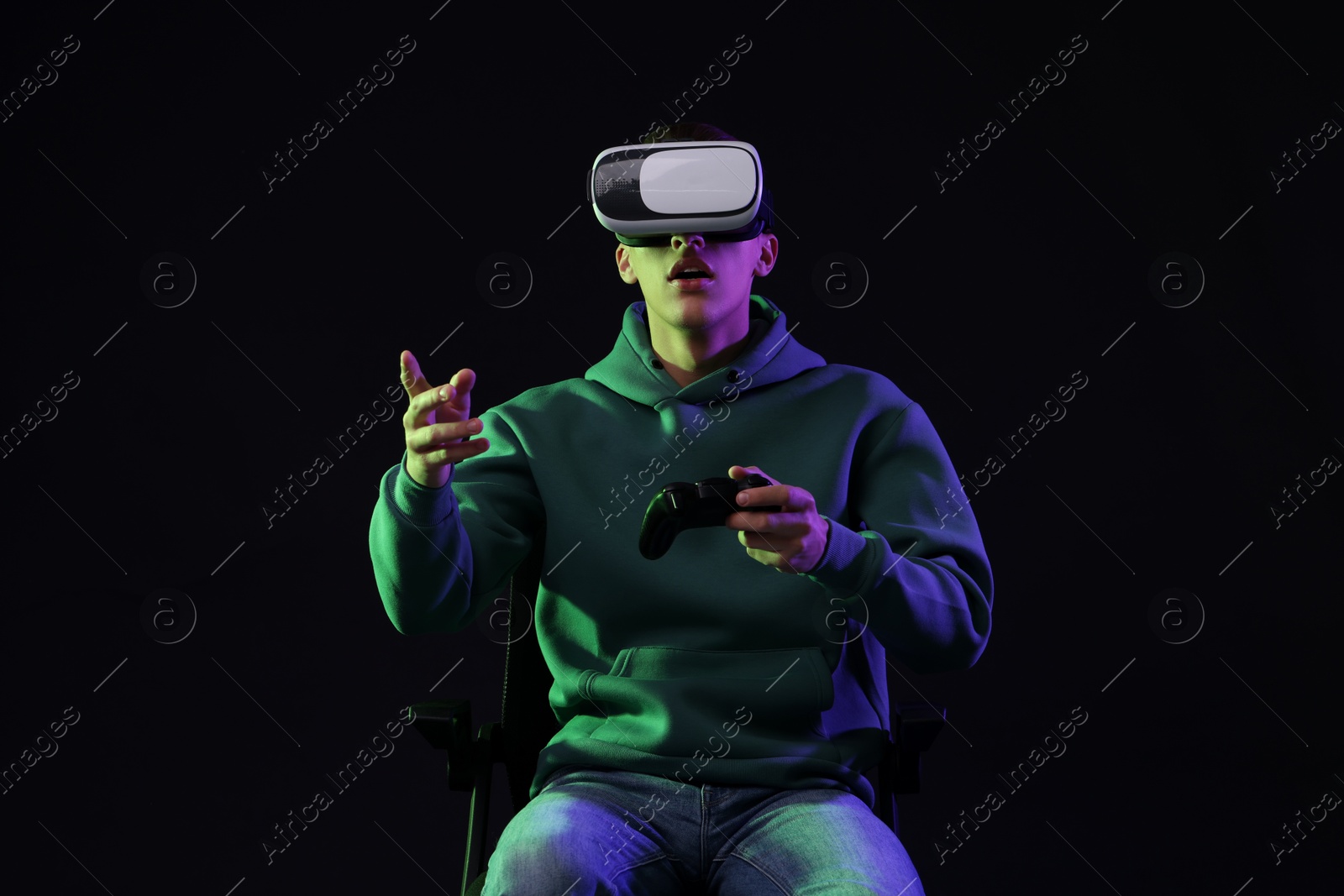 Photo of Emotional young man with virtual reality headset sitting on chair in neon lights against black background