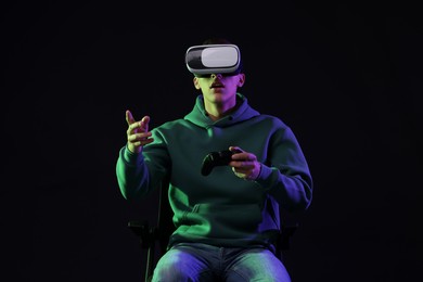 Emotional young man with virtual reality headset sitting on chair in neon lights against black background