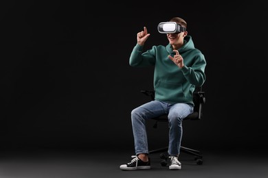 Photo of Happy young man with virtual reality headset sitting on chair against black background, space for text