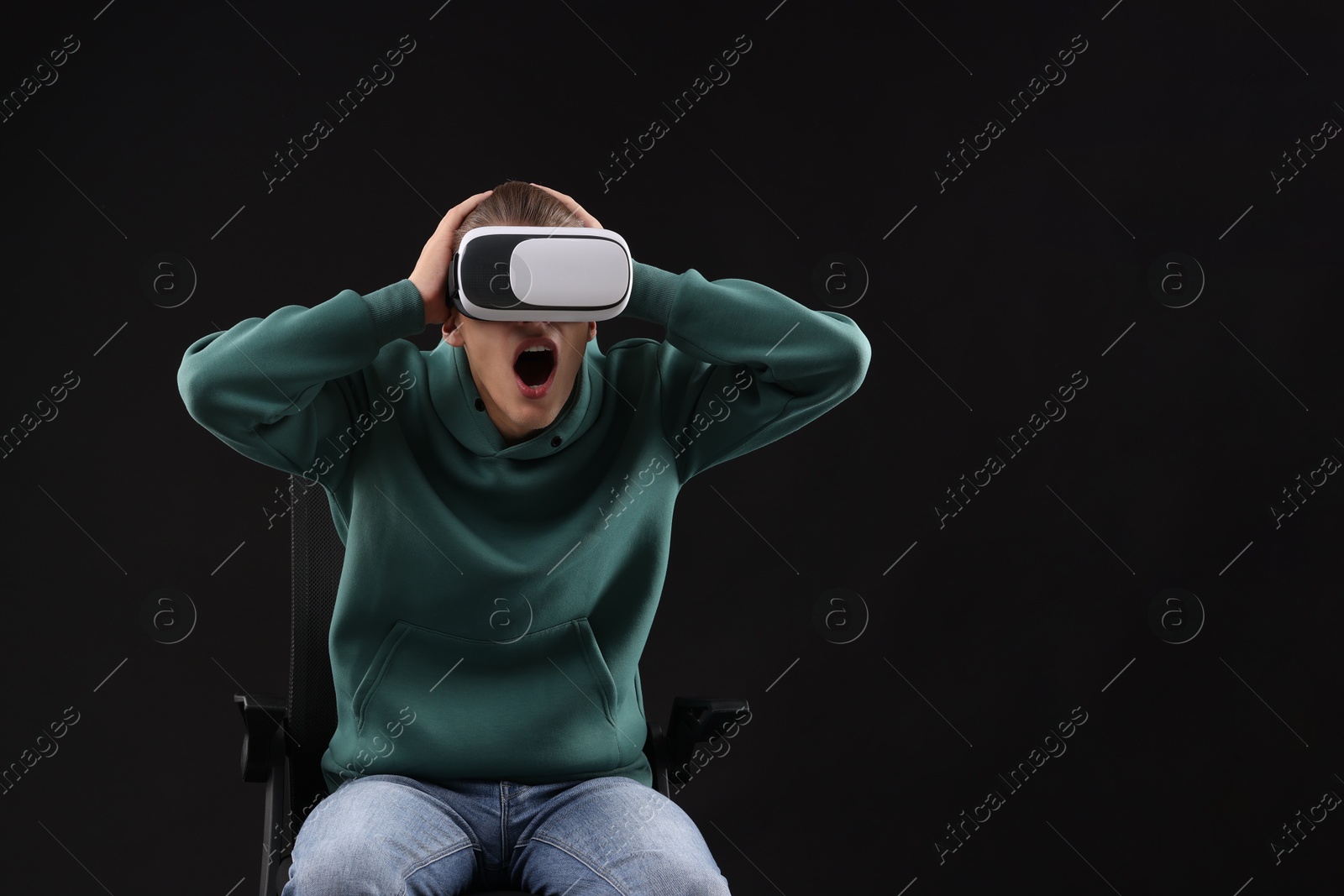 Photo of Emotional young man with virtual reality headset sitting on chair against black background, space for text