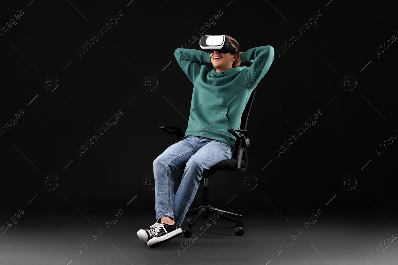 Photo of Happy young man with virtual reality headset sitting on chair against black background