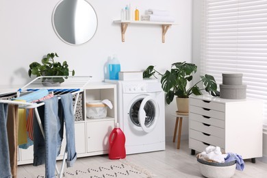 Photo of Washing machine, furniture, detergents, laundry, houseplants and drying rack indoors