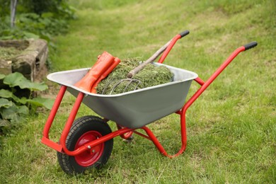 Wheelbarrow with mown grass, rubber boots and pitchfork outdoors