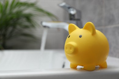 Photo of Water saving concept. Piggy bank and stream of water flowing from tap in bathroom