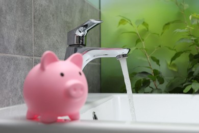 Photo of Water saving concept. Piggy bank and stream of water flowing from tap in bathroom, selective focus