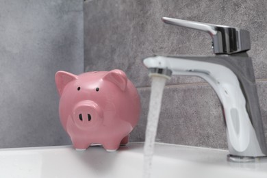 Photo of Water saving concept. Piggy bank and stream of water flowing from tap in bathroom