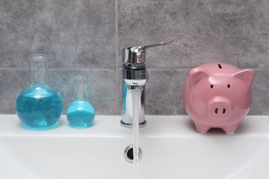 Photo of Water saving concept. Piggy bank, glassware with liquid and stream of water flowing from tap in bathroom