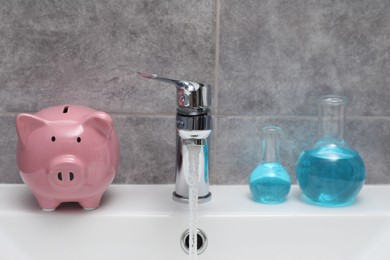 Photo of Water saving concept. Piggy bank, glassware with liquid and stream of water flowing from tap in bathroom