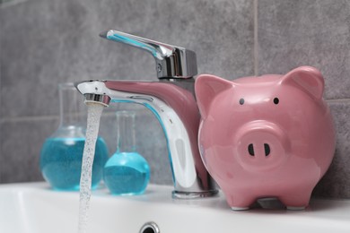 Photo of Water saving concept. Piggy bank, glassware with liquid and stream of water flowing from tap in bathroom, closeup