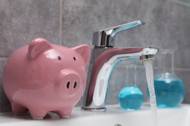 Photo of Water saving concept. Piggy bank, glassware with liquid and stream of water flowing from tap in bathroom, closeup