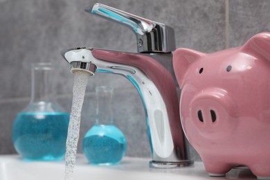 Water saving concept. Piggy bank, glassware with liquid and stream of water flowing from tap in bathroom, closeup