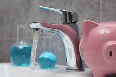 Photo of Water saving concept. Piggy bank, glassware with liquid and stream of water flowing from tap in bathroom, closeup