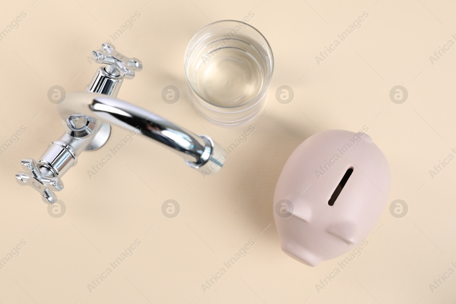 Photo of Water saving concept. Piggy bank, tap and glass of drink on pale yellow background, flat lay