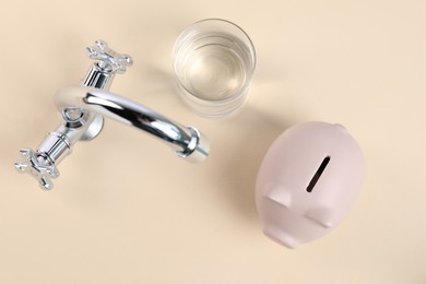 Photo of Water saving concept. Piggy bank, tap and glass of drink on pale yellow background, flat lay