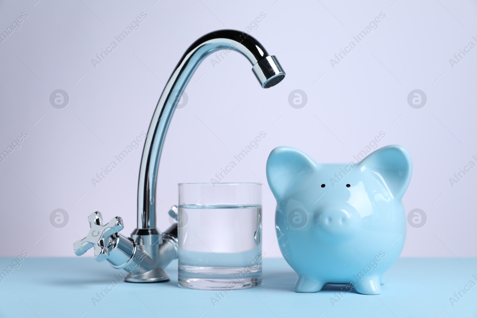 Photo of Water saving concept. Piggy bank, tap and glass of drink on light blue table against white background