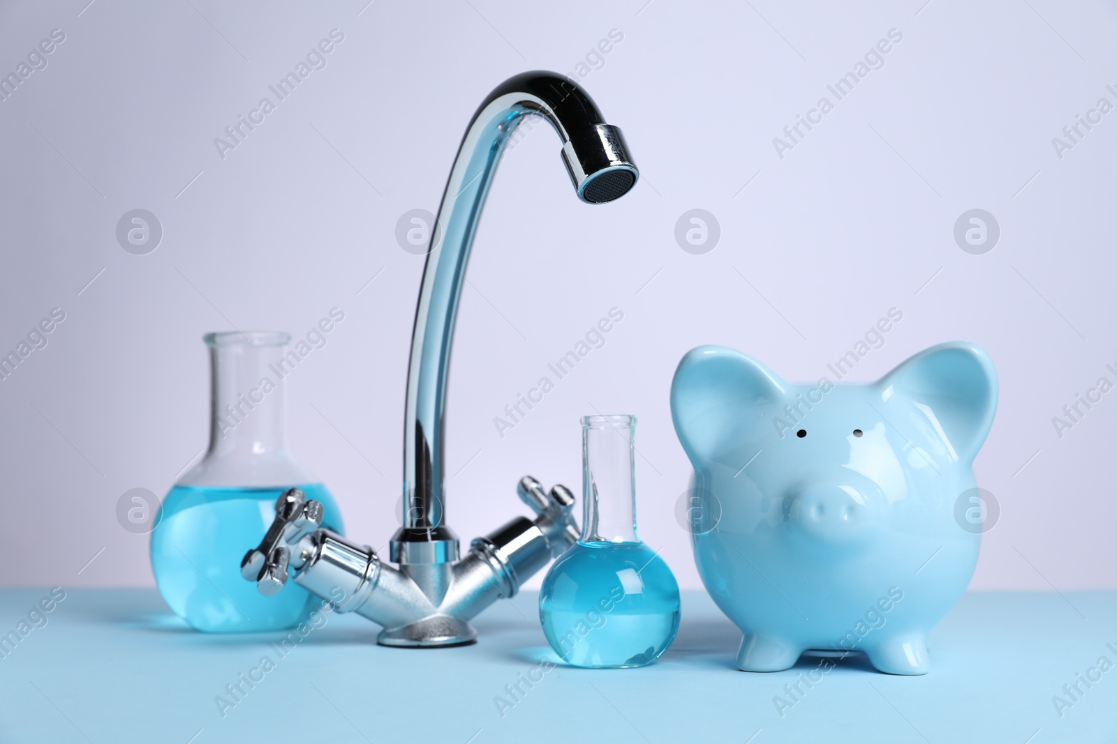 Photo of Water saving concept. Piggy bank, tap and glassware with liquid on light blue table against white background