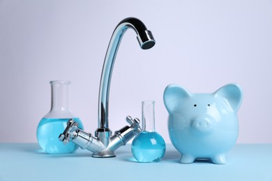 Photo of Water saving concept. Piggy bank, tap and glassware with liquid on light blue table against white background