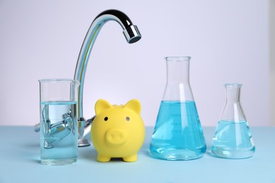 Photo of Water saving concept. Piggy bank, tap and glassware with liquid on light blue table against white background