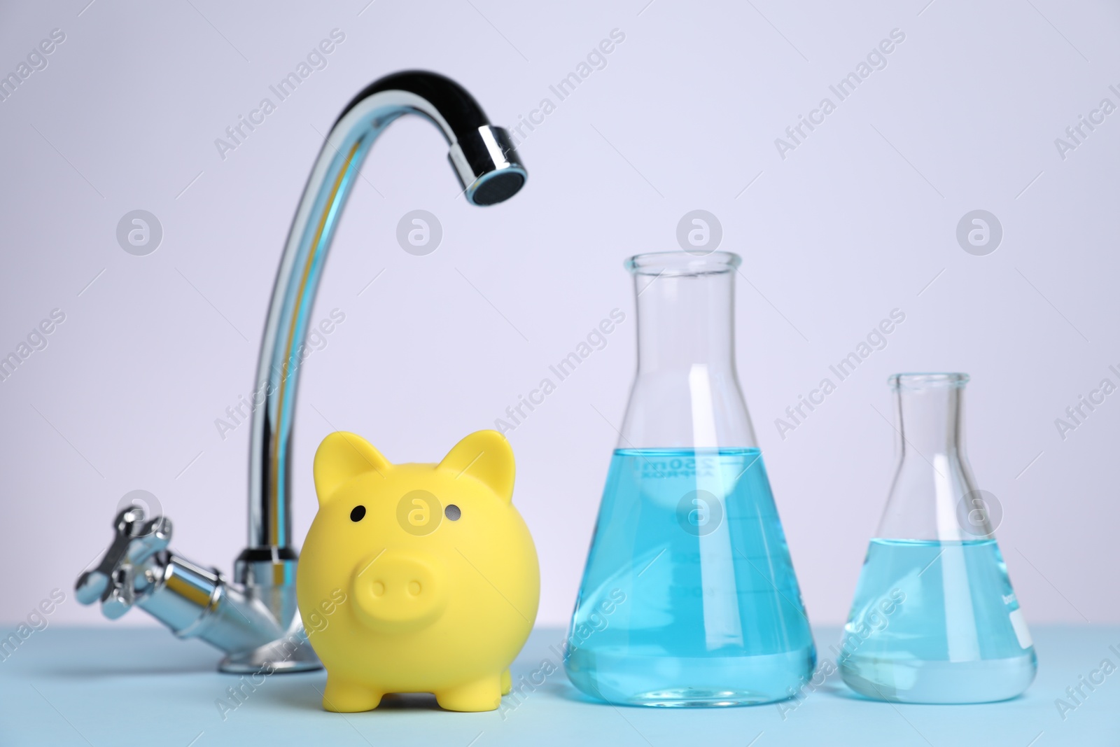 Photo of Water saving concept. Piggy bank, tap and glassware with liquid on light blue table against white background