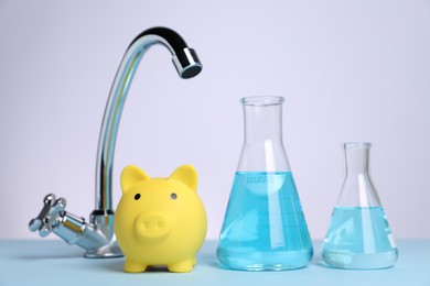 Photo of Water saving concept. Piggy bank, tap and glassware with liquid on light blue table against white background