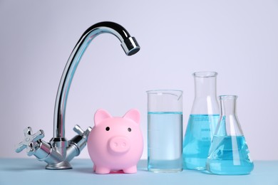 Water saving concept. Piggy bank, tap and glassware with liquid on light blue table against white background