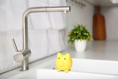 Photo of Water saving concept. Stream of water flowing from tap onto piggy bank in kitchen