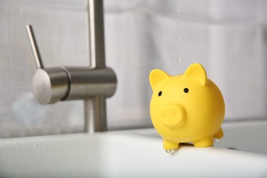 Photo of Water saving concept. Stream of water flowing from tap onto piggy bank in kitchen