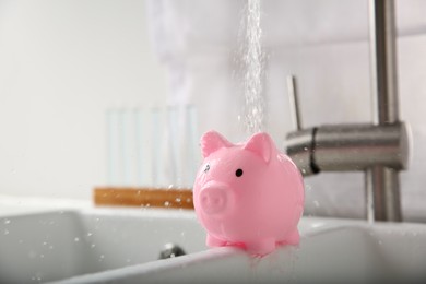 Photo of Water saving concept. Stream of water flowing from tap onto piggy bank in kitchen