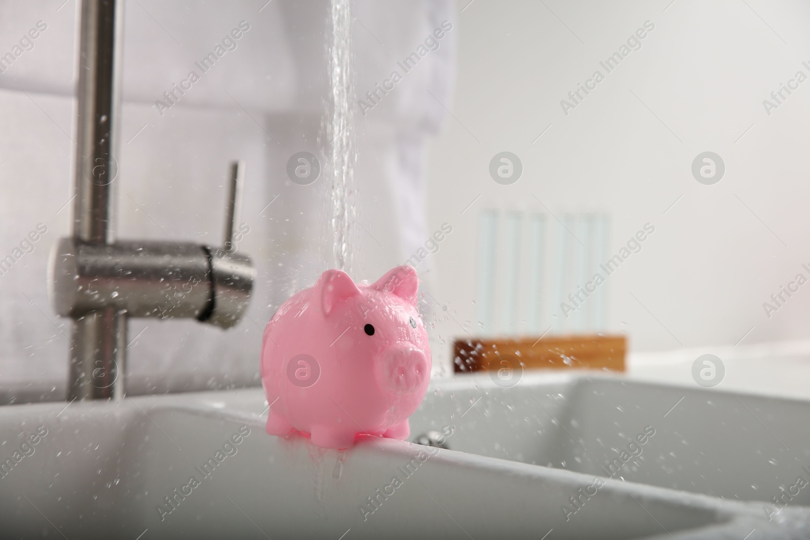 Water saving concept. Stream of water flowing from tap onto piggy bank in kitchen Photo of Water saving concept. Stream of water flowing from tap onto piggy bank in kitchen