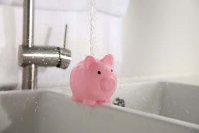 Photo of Water saving concept. Stream of water flowing from tap onto piggy bank in kitchen