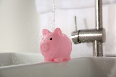 Photo of Water saving concept. Stream of water flowing from tap onto piggy bank in kitchen