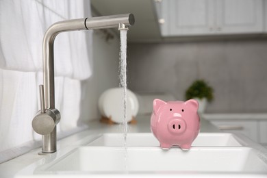 Photo of Water saving concept. Piggy bank and stream of water flowing from tap in kitchen