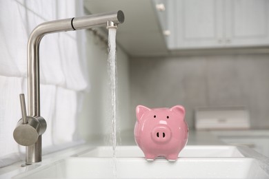 Photo of Water saving concept. Piggy bank and stream of water flowing from tap in kitchen