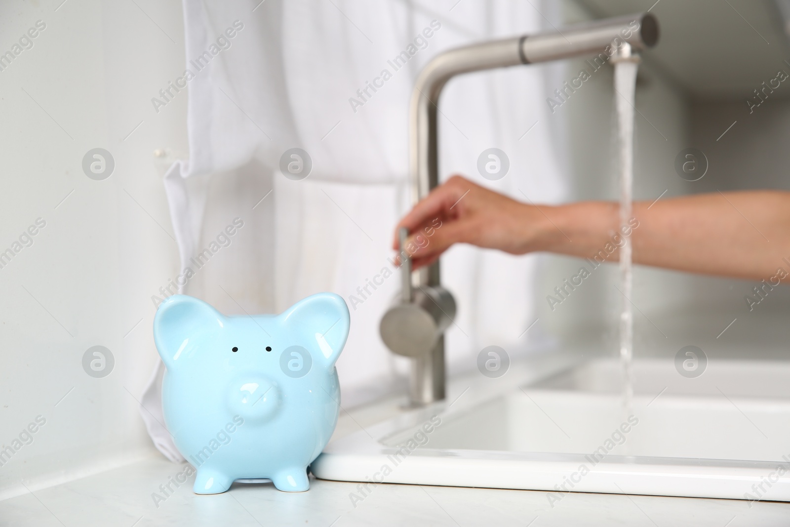 Photo of Water saving concept. Woman checking water flow in kitchen, focus on piggy bank